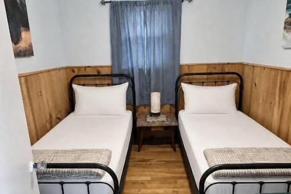 Une chambre avec deux lits simples, un ventilateur de plafond, une petite table avec une bougie et une fenêtre avec des rideaux.