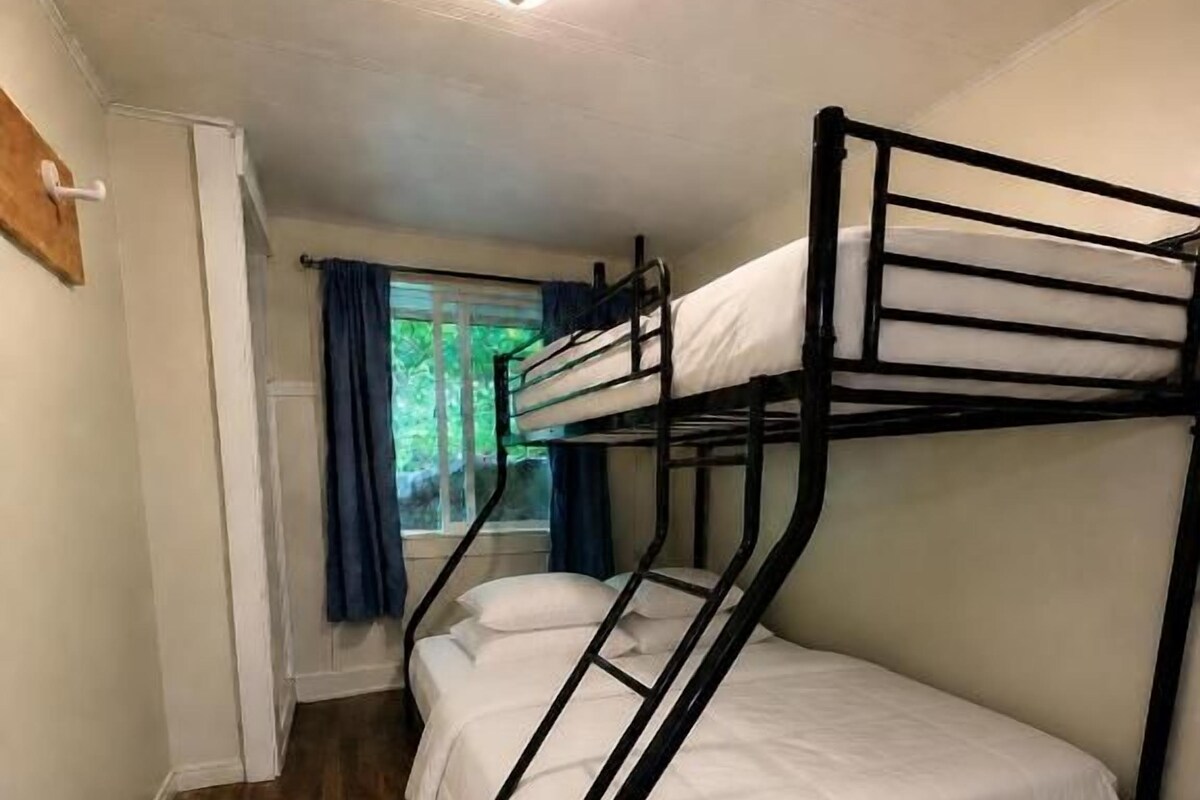 Un lit superposé avec un matelas blanc et une structure en métal noir, dans une pièce dont le sol est en bois et qui possède une fenêtre avec des rideaux.