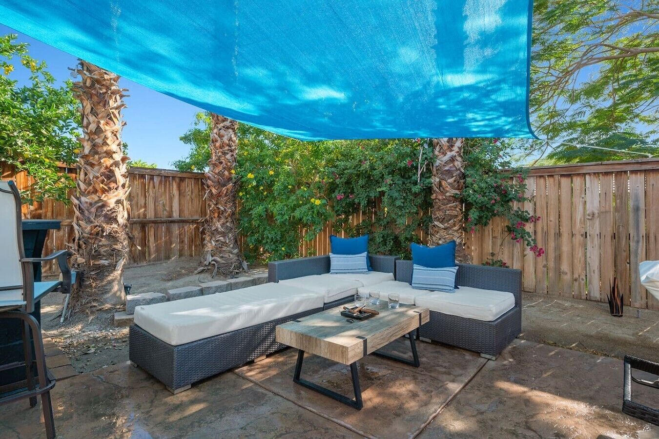 Eine Terrasse mit einem großen, blauen Sonnensegel, Korbmöbeln und einem Holztisch.