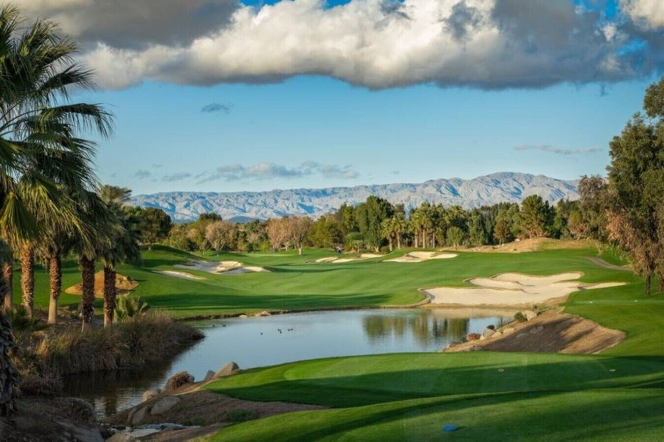 Un parcours de golf avec un étang, des palmiers et des montagnes en arrière-plan.
