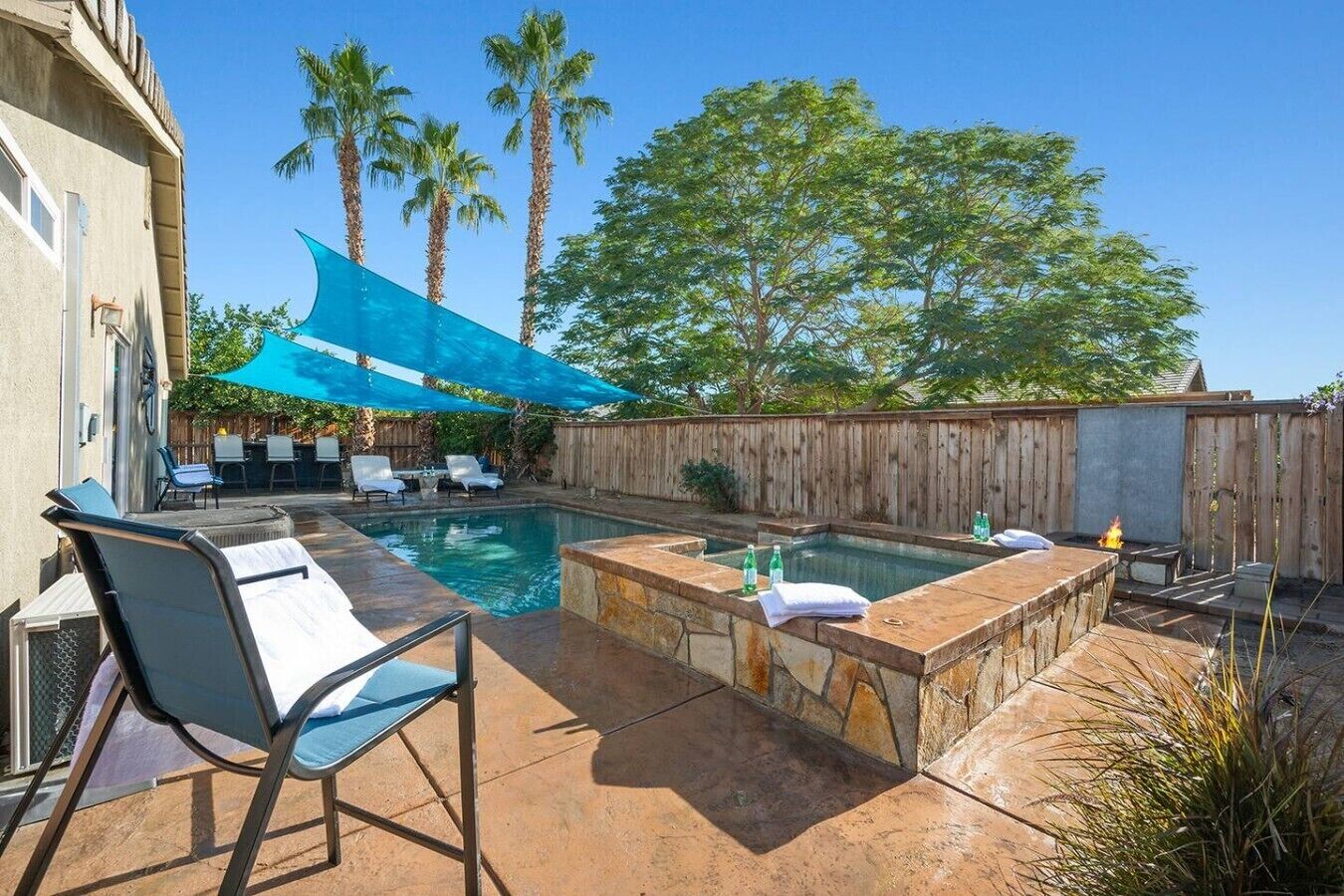 A backyard with a swimming pool, a hot tub, and a patio area with chairs and a table.