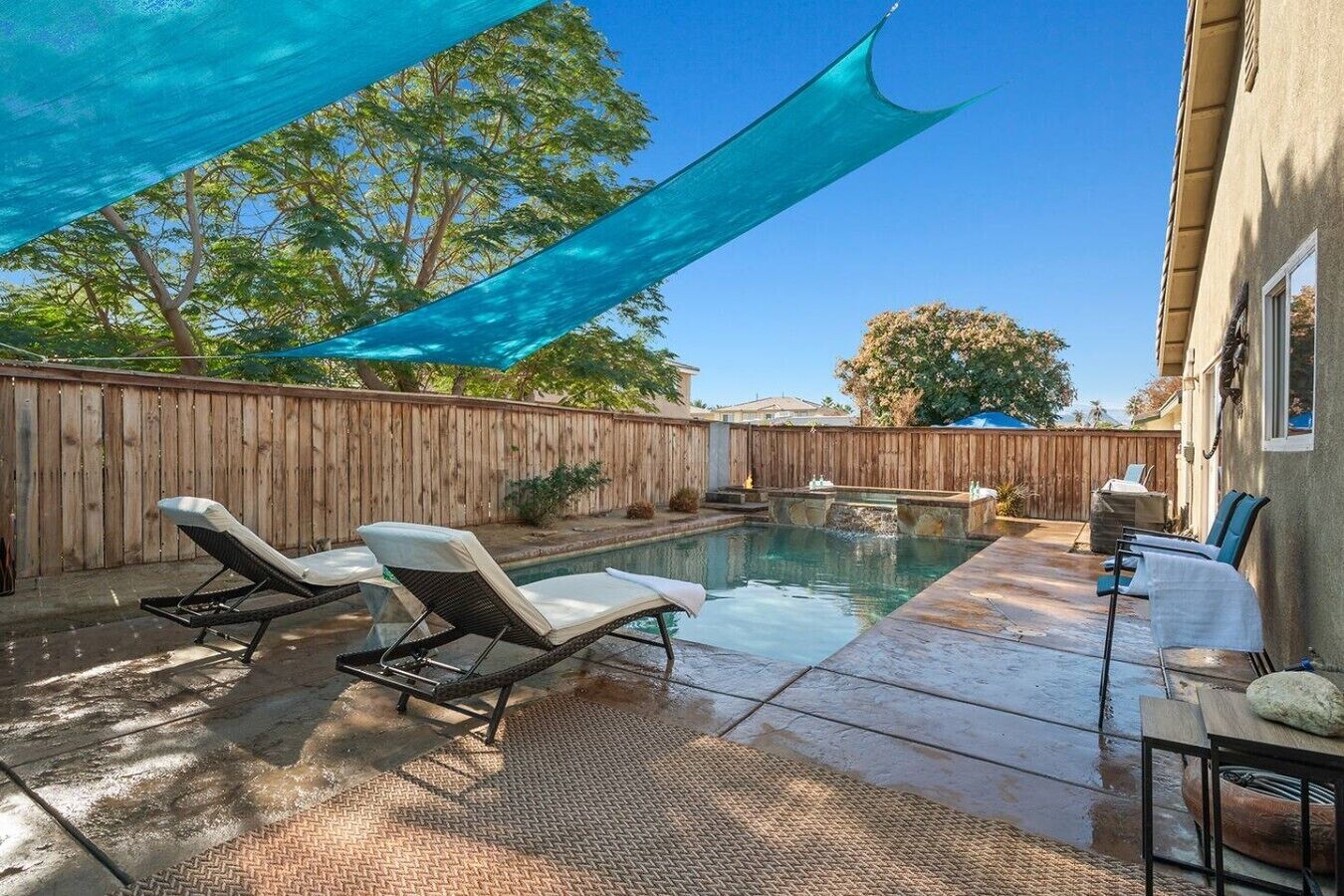 A backyard with a swimming pool, two lounge chairs, a wooden fence, and a shaded area.