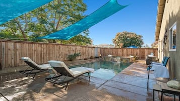 A backyard with a swimming pool, two lounge chairs, a wooden fence, and a shaded area.
