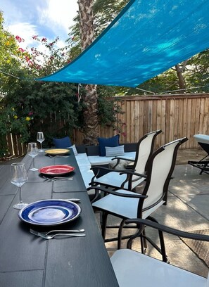 Un patio exterior con mesa lista para comer, sillas y un toldo azul por encima.