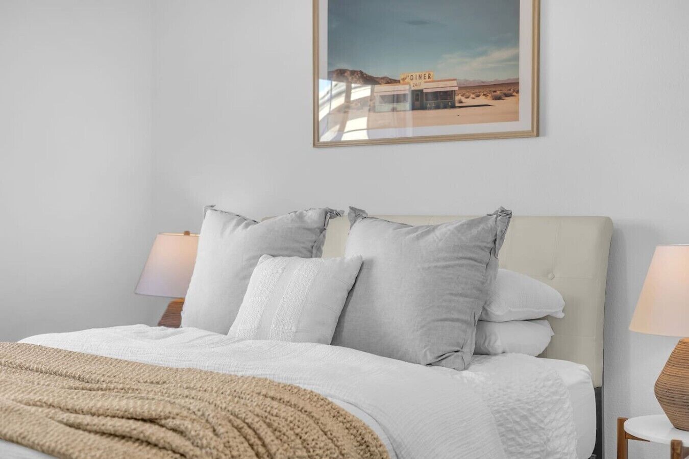 Ein Bett mit weißer Decke und Kissen, einer beige farbenen Tagesdecke und einem gerahmten Bild von einer Diner-Kneipe an der Wand.