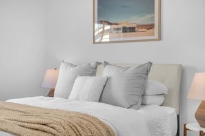 A bed with a white comforter and pillows, a beige throw blanket, and a framed picture of a diner on the wall. - Early Check-In. AC Comfort + Pool, Spa +FAST WIFI (Coachella)