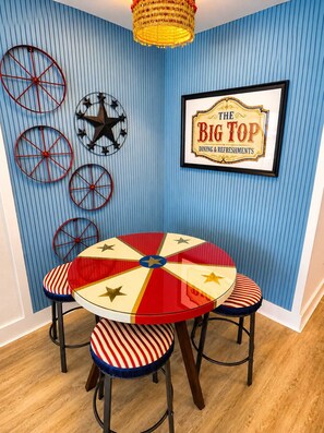 A dining area with a round table and four chairs, a wall with vertical blue stripes, and a sign that reads 'THE BIG TOP DINING & REFRESHMENTS'. - Circus-Themed Oceanfront Condo! Yachtsman 1001 (Myrtle Beach)