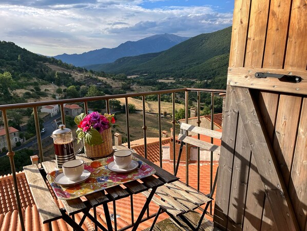 Een balkon met een tafeltje voor twee, een koffiepot, een mand met bloemen en uitzicht op een bergachtig landschap.