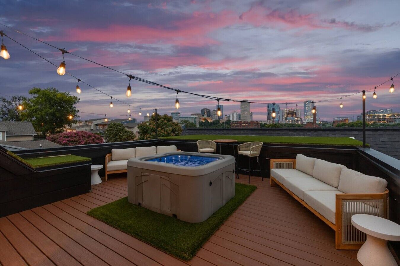A rooftop deck with a hot tub, seating area, and string lights against a city skyline at dusk.