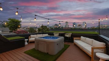 A rooftop deck with a hot tub, seating area, and string lights against a city skyline at dusk.