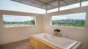 A bathroom with a large bathtub, two windows with views of a landscape, and a shower area. - Kayun Retreat Mas Suite (Ubud)