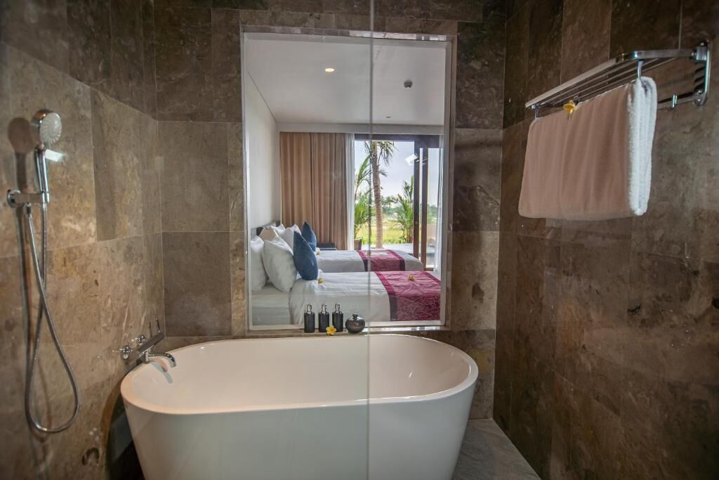 A modern bathroom with a freestanding bathtub, a shower, and a large mirror reflecting a bedroom with a bed and curtains.