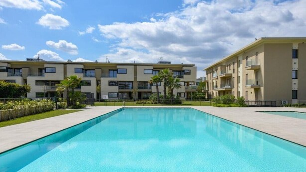 A swimming pool with a clear blue water surface, surrounded by a low-rise apartment building with balconies. - Oasi del Garda in Desenzano - Pool, Garden, and Garage (Desenzano del Garda)