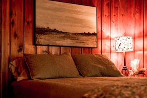 En säng med två kuddar, en inramad landskapsmålning ovanför och en lampa med ett blommönster på nattduksbordet.