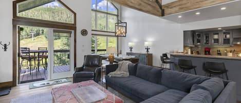 A spacious living area with a large gray sofa, a leather chair, and a dining table with chairs. There are large windows offering a view of a forested area.
