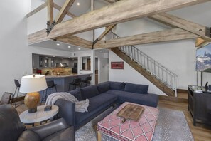 A modern living room with a large L-shaped sectional sofa, a round coffee table, and a lamp. A staircase with a metal railing leads to an upper level. - Mountain Village Chalet | Ski-In/Out+ Hot Tub (Mountain Village)