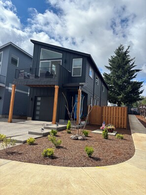 A modern two-story house with a balcony, a driveway, and a well-maintained garden with shrubs and a small tree. - YOUR MT HOOD GATEWAY STARTS HERE! NEW 3 BD 2.5 BA HOME W/SMALL TOWN CONVENIENCE! (Sandy)