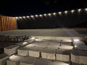 Een terras in de open lucht 's nachts, met een betonnen pad, houten omheining en lichtslingers.