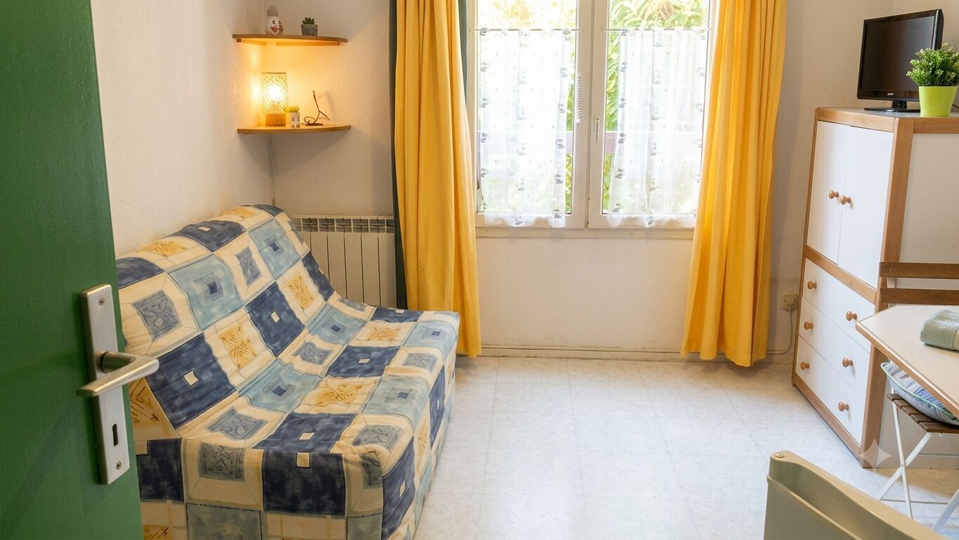 Ein Zimmer mit einer gemusterten Couch, einem Schrank mit Schubladen, einem Fernseher auf einem Beistelltisch und einem Fenster mit Vorhängen.