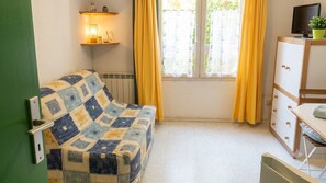 A room with a patterned sofa, a cabinet with drawers, a television on a stand, and a window with curtains. - Quiet Studio in Lamalou – 10 min from Thermal Baths – Wi-Fi – Pets Welcome (Lamalou-les-Bains)