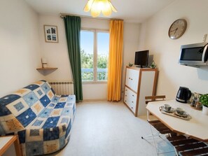 A room with a sofa, a television, a table with cups and a kettle, a clock, and a window with curtains. - Quiet Studio in Lamalou – 10 min from Thermal Baths – Wi-Fi – Pets Welcome (Lamalou-les-Bains)