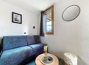 A room with a dark blue sofa, a small round table with a vase, a framed picture on the wall, and a window with curtains. - La Plagne Montalbert, ski-in/ski-out, in the heart of the resort, unobstructed view, ski lift. (Aime-la-Plagne)