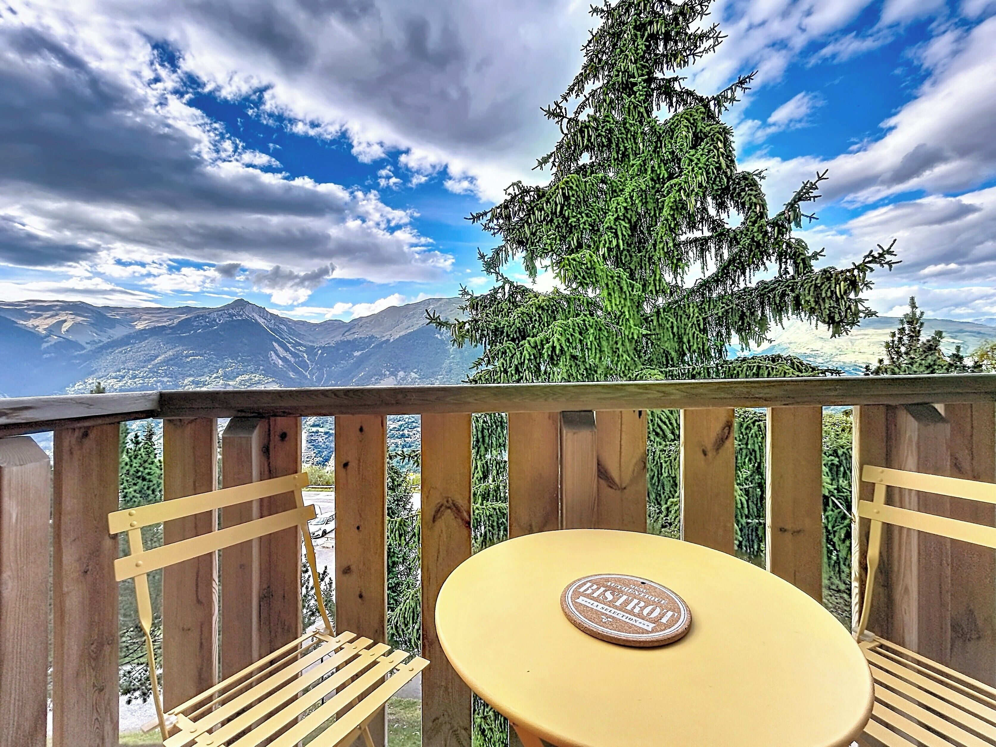 A wooden deck with a round table and a bench, overlooking a mountainous landscape with a clear sky.