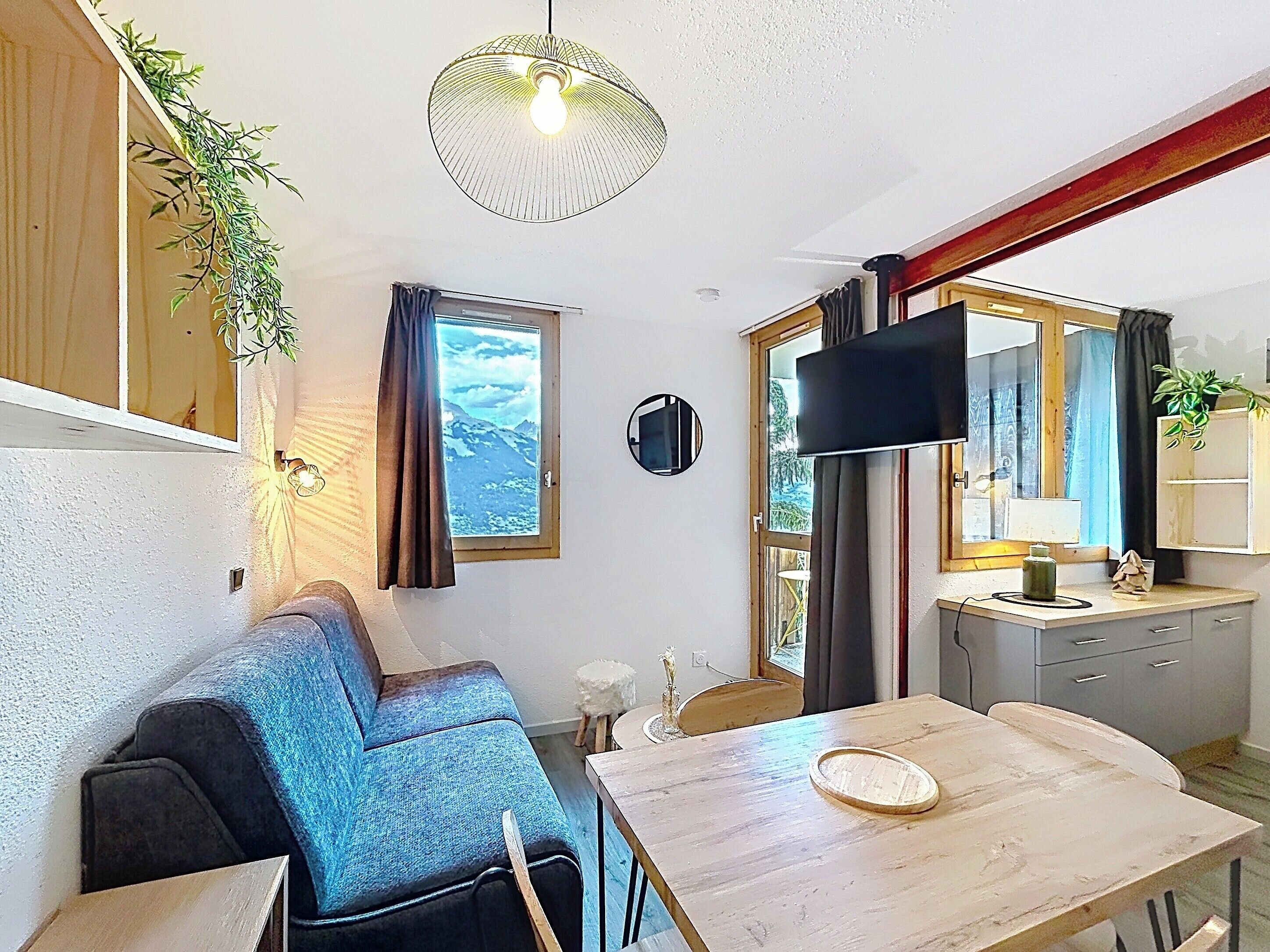 A compact living space with a sofa, a dining table, a TV, and a window with a view of mountains.
