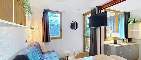 Ein kompakter Wohnraum mit einer Couch, einem Esstisch, einem Fernseher und einem Fenster mit Bergblick.