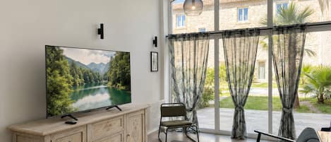 En stue med et stort fladskærms-tv, et træskab, et sort sofabord, en grå sofa og udsigt til en stengbygning gennem store vinduer.