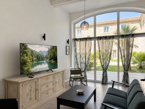 Een woonkamer met een grote flatscreen televisie, een houten kast, een zwarte salontafel, een grijze bank en uitzicht op een stenen gebouw door grote ramen.
