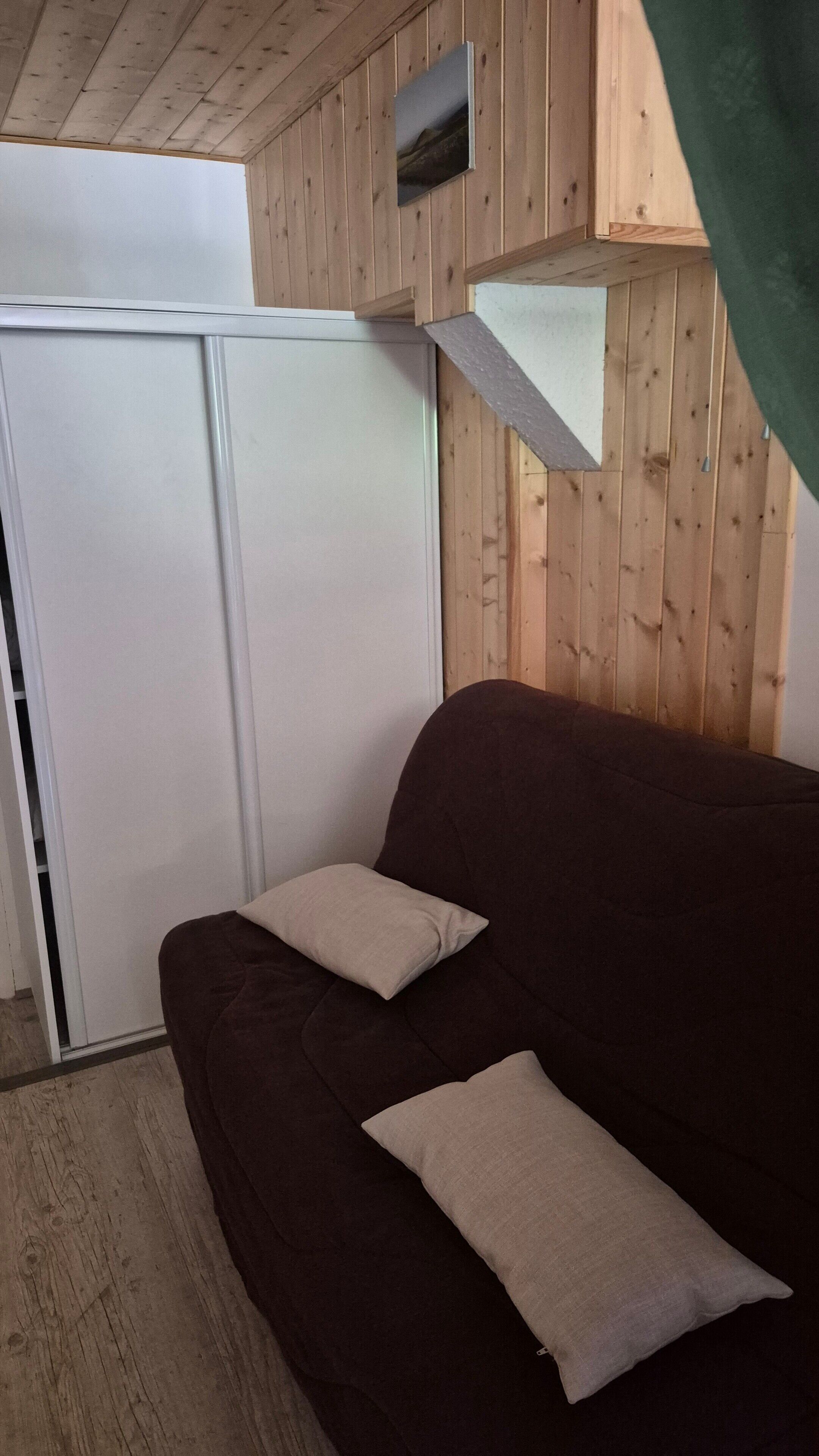 A room with a dark brown sofa, two light-colored pillows, a white wardrobe, and wooden panel walls.