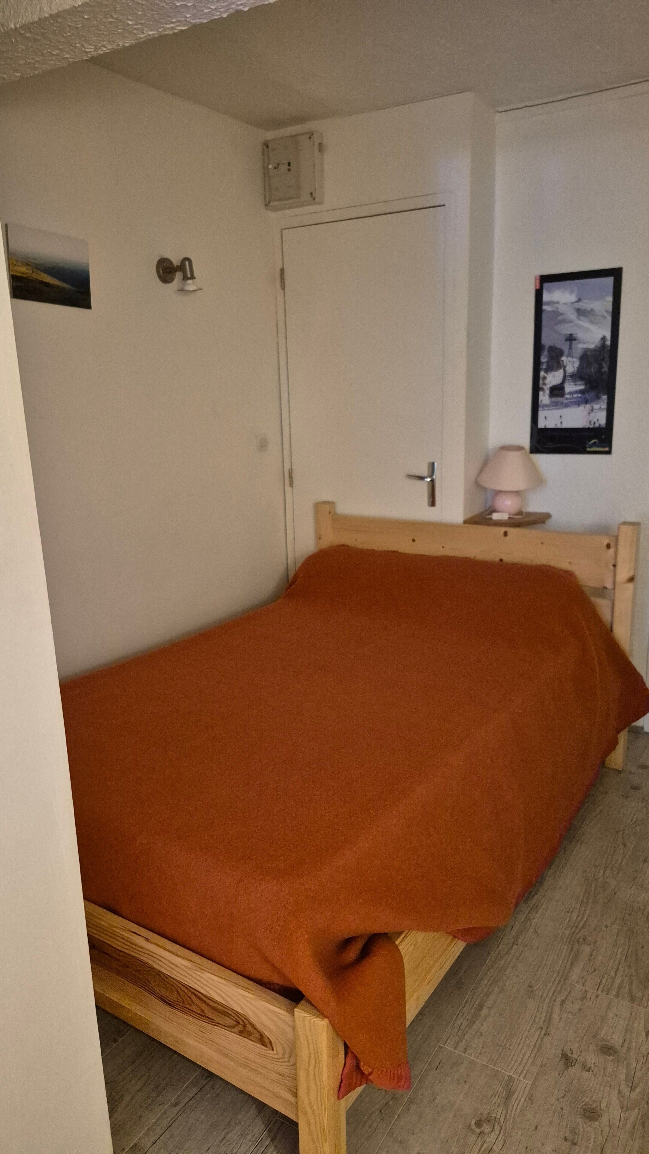 En enkel seng med et brunt tæppe, et træhovedgærde, en hvid dør, et lille bord med en lampe og et billede i ramme på væggen.