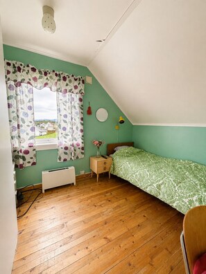 Ein Schlafzimmer mit schrägverdeckter Decke, einem Heizkörper, einem Nachttisch mit Lampe und einem Fenster mit blumigem Vorhang.