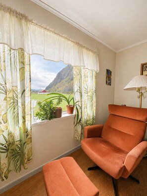 A room with a view of a mountainous landscape, a window with curtains, a chair, a table, and a lamp. - Midnight Sun House in Eggum, Lofoten (Vestvågøy)