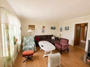 Ein Wohnzimmer mit einer Couch, einem Sessel, einem Hocker und einem Tisch. An den Wänden sind Wandlampen, ein Heizkörper und gerahmte Bilder angebracht.
