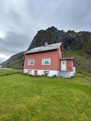 A red wooden house with a white door and windows, set against a mountainous backdrop. - Midnight Sun House in Eggum, Lofoten (Vestvågøy)