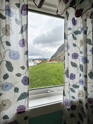Ein Fenster mit Vorhängen mit Blumenmuster, Blick auf ein grünes Feld mit Gebäuden und einem bergigen Hintergrund.