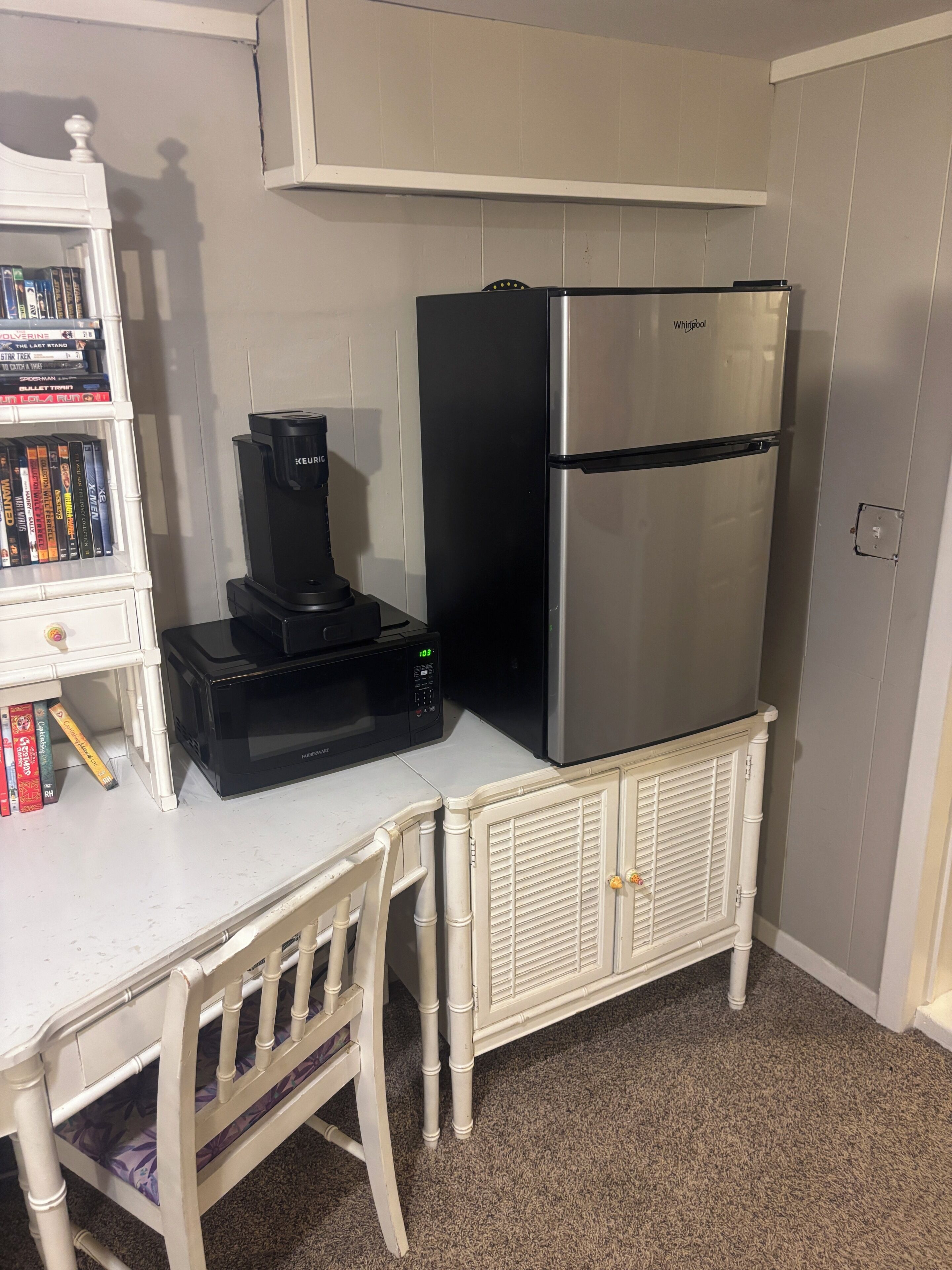 Eine kleine Küchenzeile mit weißer Arbeitsplatte, einer schwarzen Kaffeemaschine, einem Edelstahlkühlschrank und einem weißen Schrank mit Büchern.