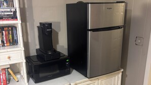 A compact kitchenette with a white countertop, a black coffee maker, a stainless steel refrigerator, and a white cabinet with books.