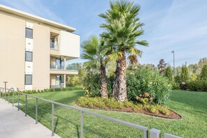 Ein modernes Gebäude mit flachem Dach, einem Balkon und einem gepflegten Garten mit Palmen und Sträuchern.