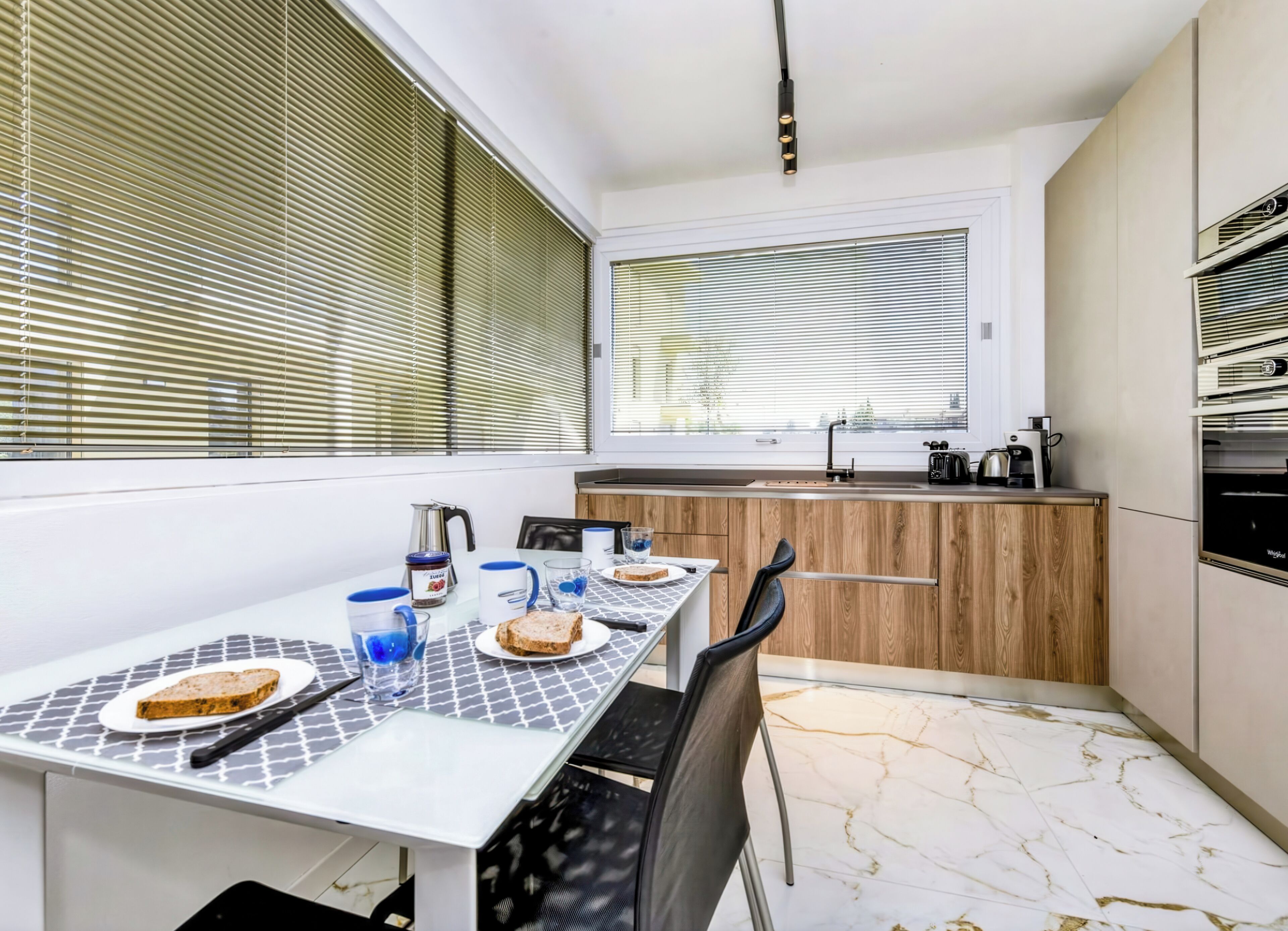 A modern kitchen with a dining area, featuring a marble floor, wooden cabinets, and a window with blinds.