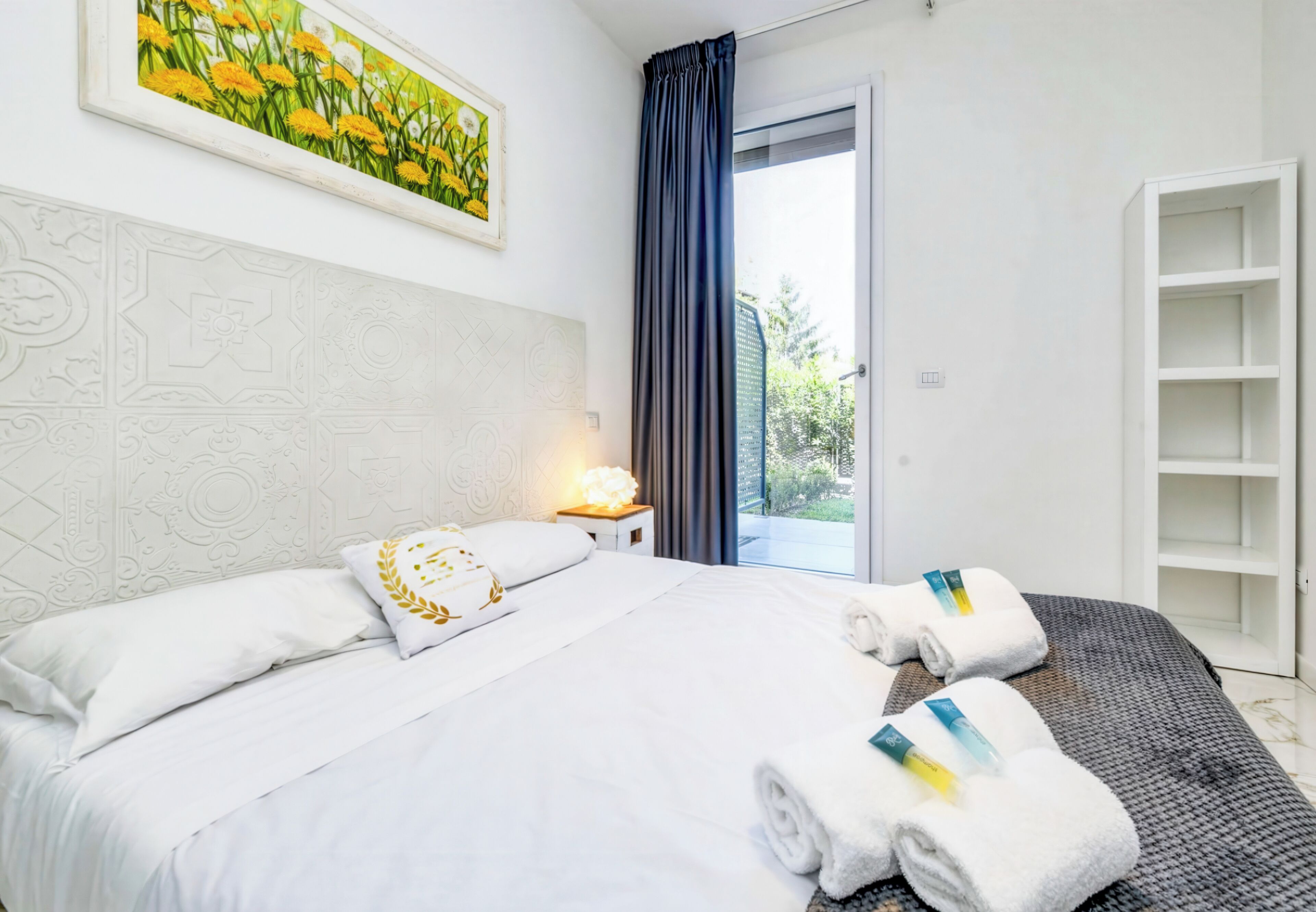 A hotel room with a bed, a nightstand, a painting, a window with curtains, and a built-in shelving unit.