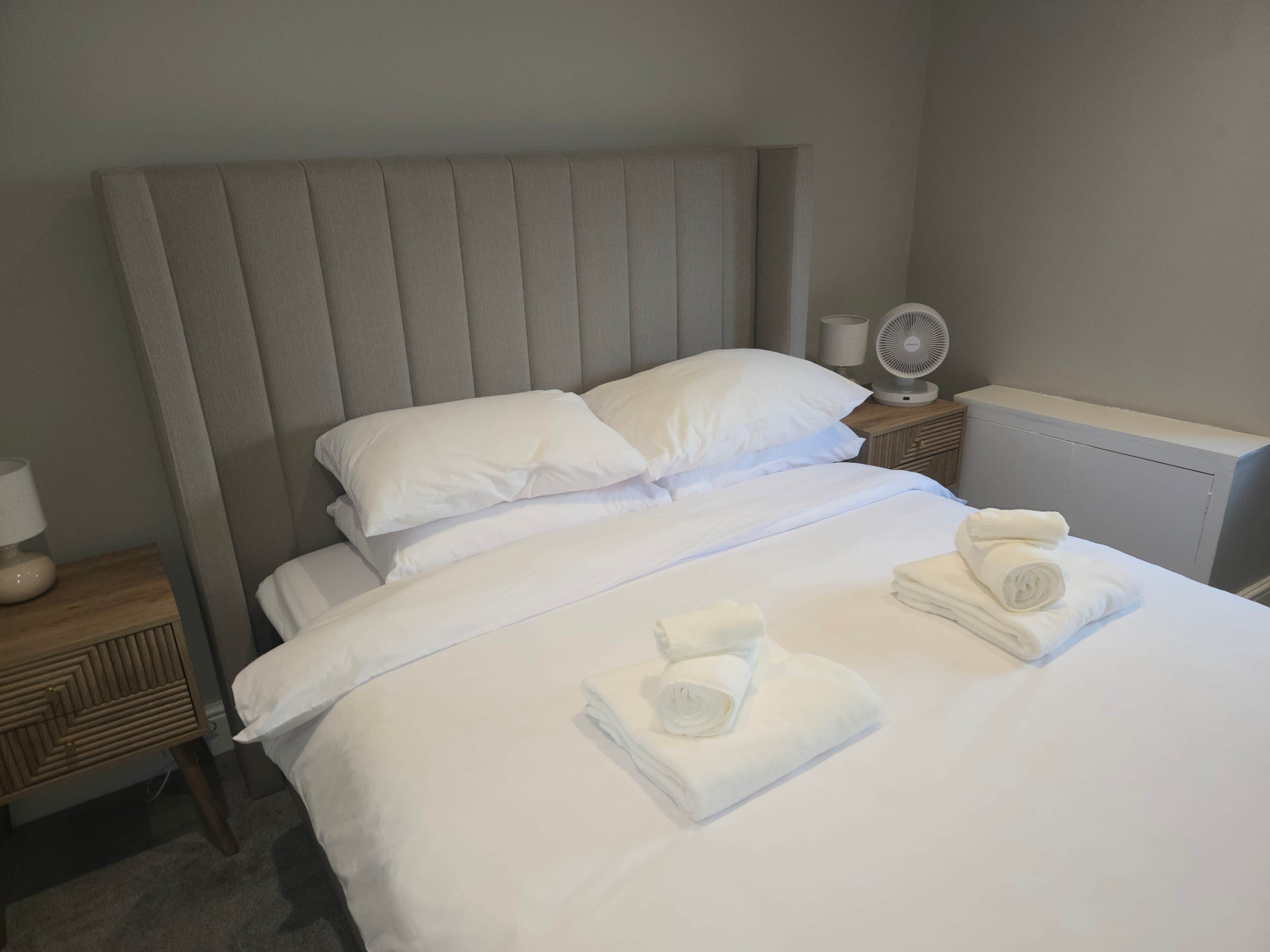 Una cama bien tendida con sábanas blancas, un cabecero y dos mesitas de noche con lámparas y un ventilador.