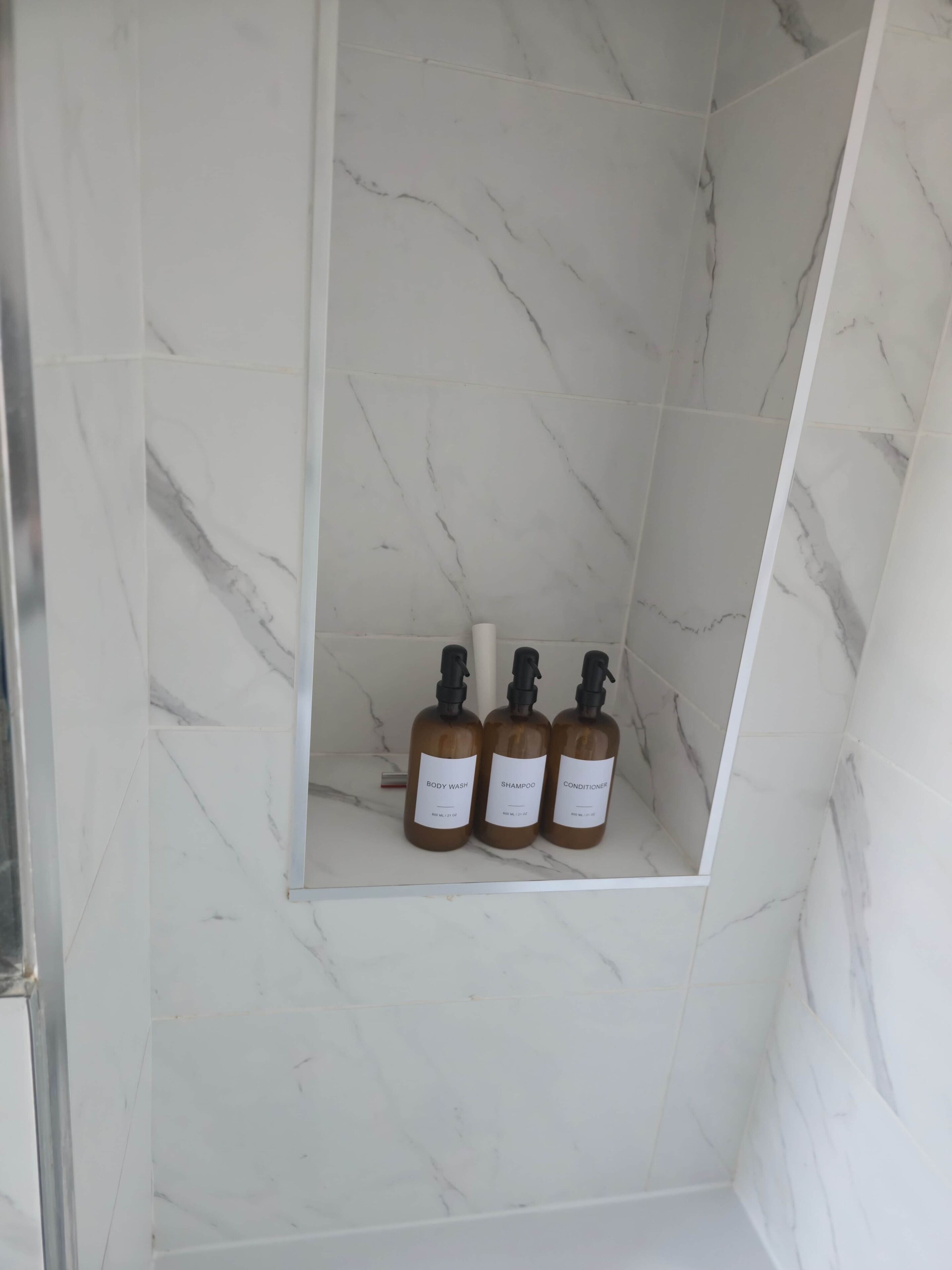 A bathroom with a marble wall and a shelf holding three bottles of body wash, shampoo, and conditioner.
