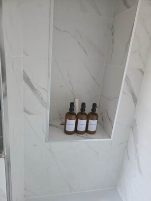 A bathroom with a marble wall and a shelf holding three bottles of body wash, shampoo, and conditioner. - Stylish Brighton Flat Near Station, Beach & Lanes (Brighton and Hove)