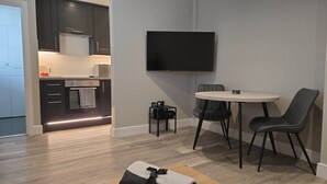 Eine moderne Küche mit dunklen Schränken, einem eingebauten Backofen und einer Mikrowelle. Ein Essbereich mit rundem Tisch und zwei Stühlen sowie ein flachbildfernseher an der Wand sind vorhanden.
