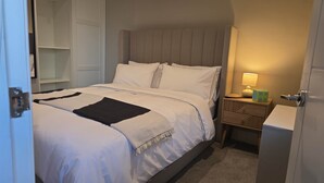 Ein ordentlich bezogenes Bett mit Kopfteil, ein Nachttisch mit Lampe und ein kleiner Beistelltisch mit Lautsprecher.
