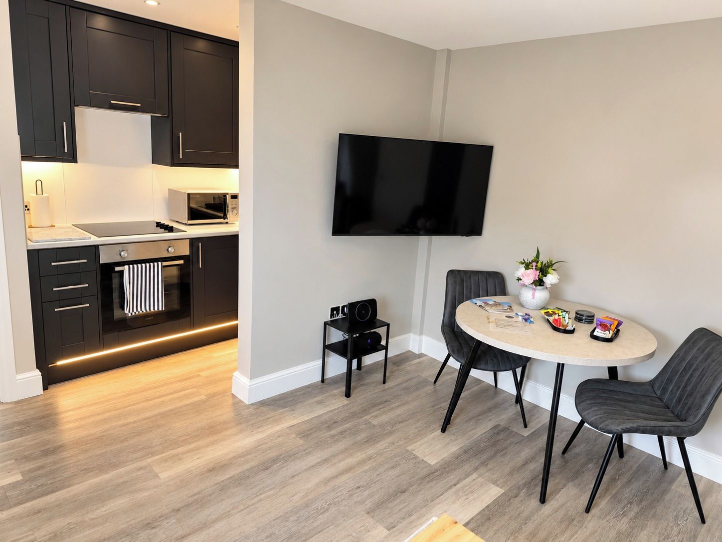 A modern kitchen with dark cabinetry, a microwave, and a stove. There is a dining area with a round table, two chairs, and a flat-screen TV mounted on the wall.