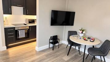 A modern kitchen with dark cabinetry, a microwave, and a stove. There is a dining area with a round table, two chairs, and a flat-screen TV mounted on the wall.